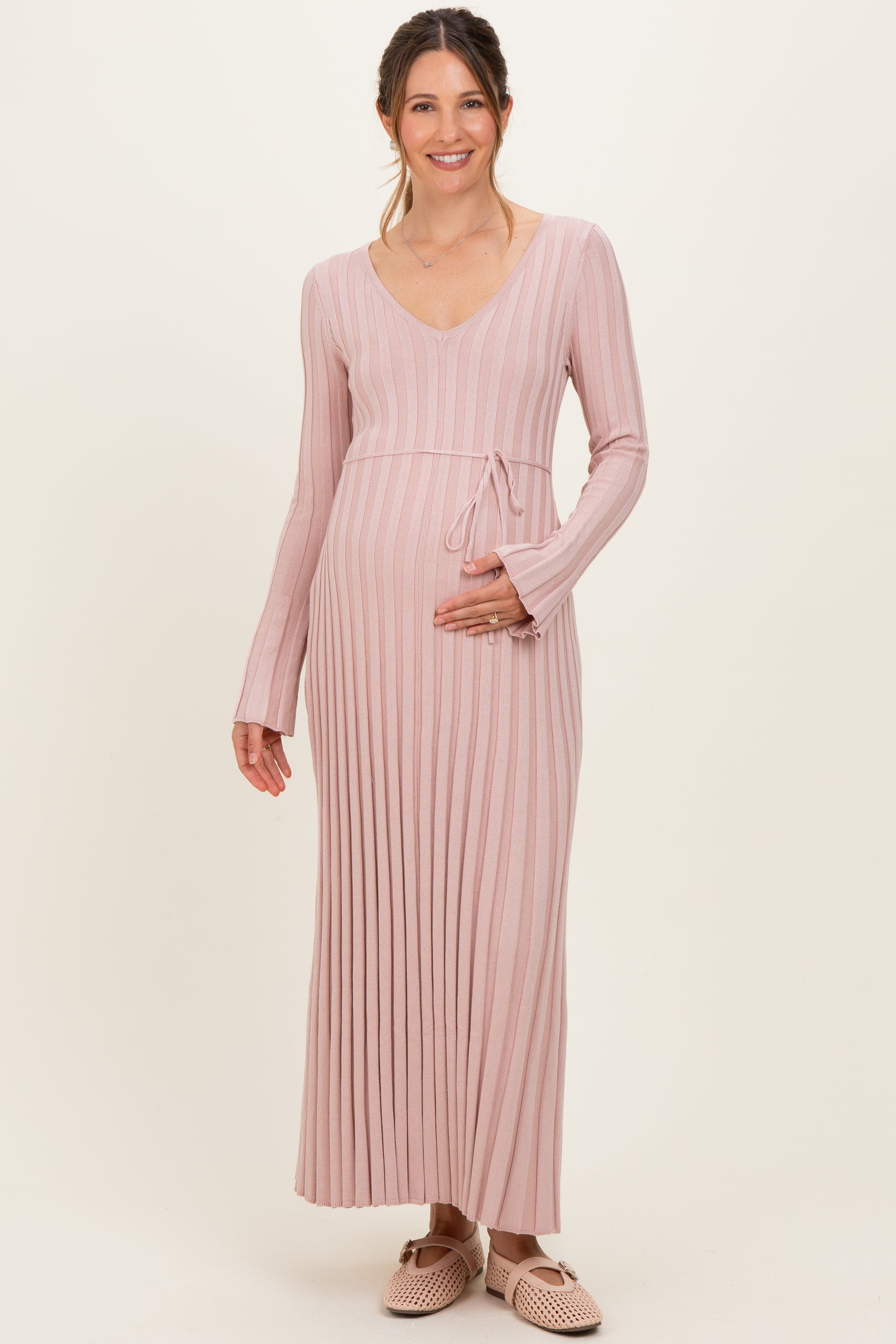 woman wearing a light pink ribbed maternity dress with long sleeves and a tie waist, holding her baby bump