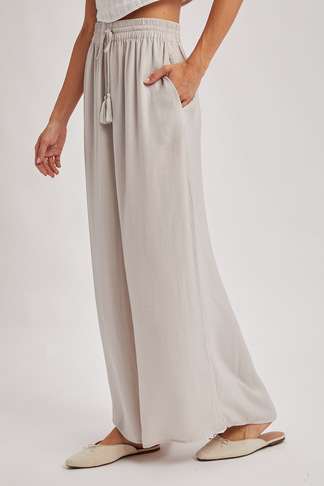 woman wearing light gray wide-leg pants with a drawstring and tassel detail, featuring side pockets