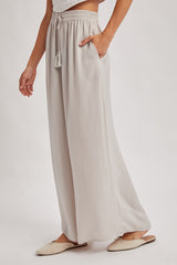 woman wearing light gray wide-leg pants with a drawstring and tassel detail, featuring side pockets
