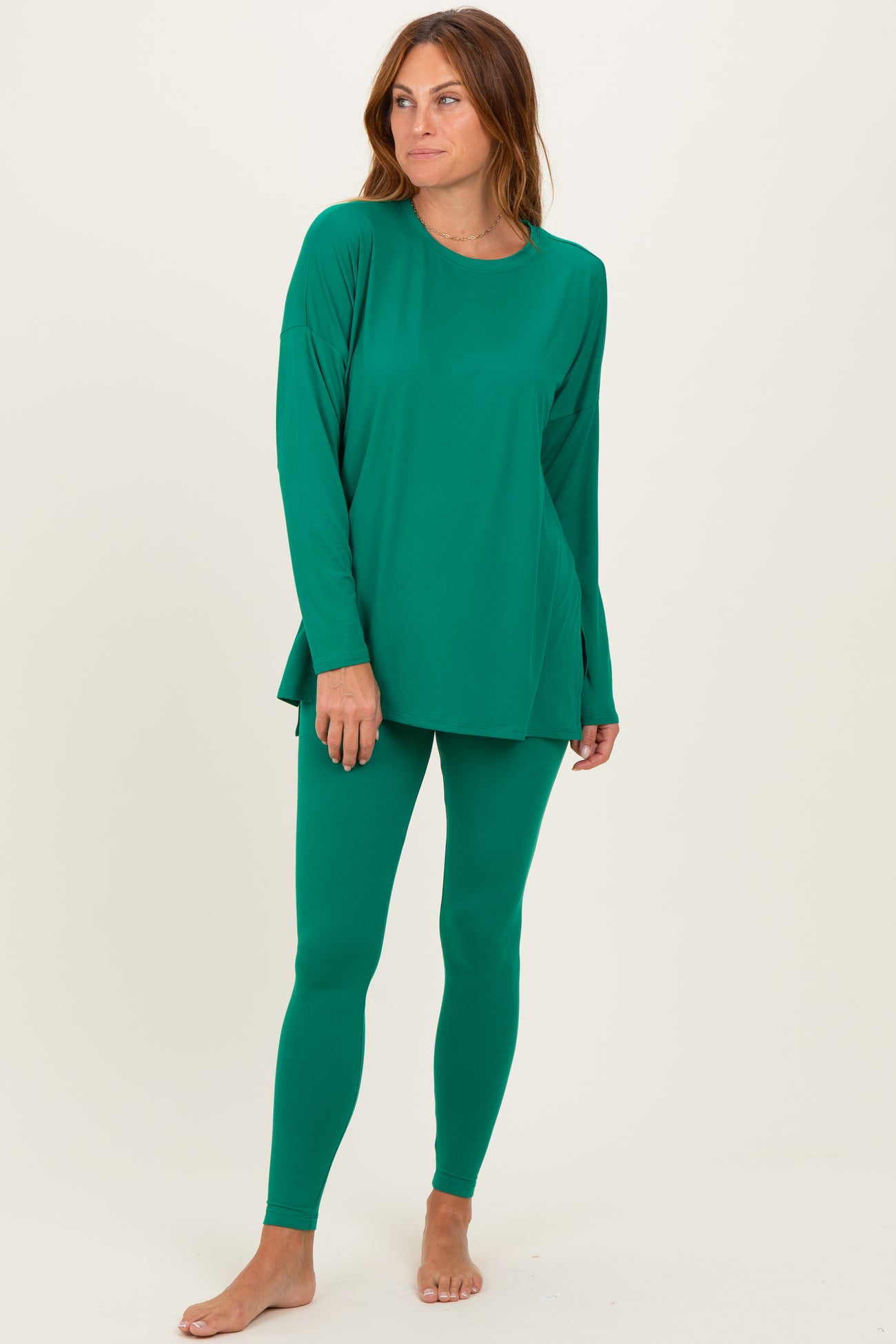 woman wearing a green long sleeve top and matching leggings, standing barefoot against a neutral background