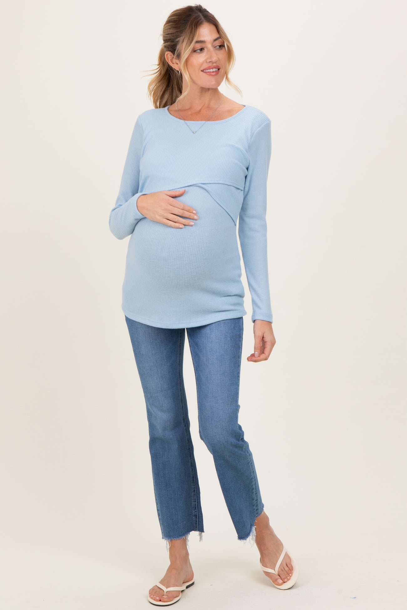 woman wearing a light blue long sleeve maternity top with gathered detail and holding her baby bump, paired with flared jeans