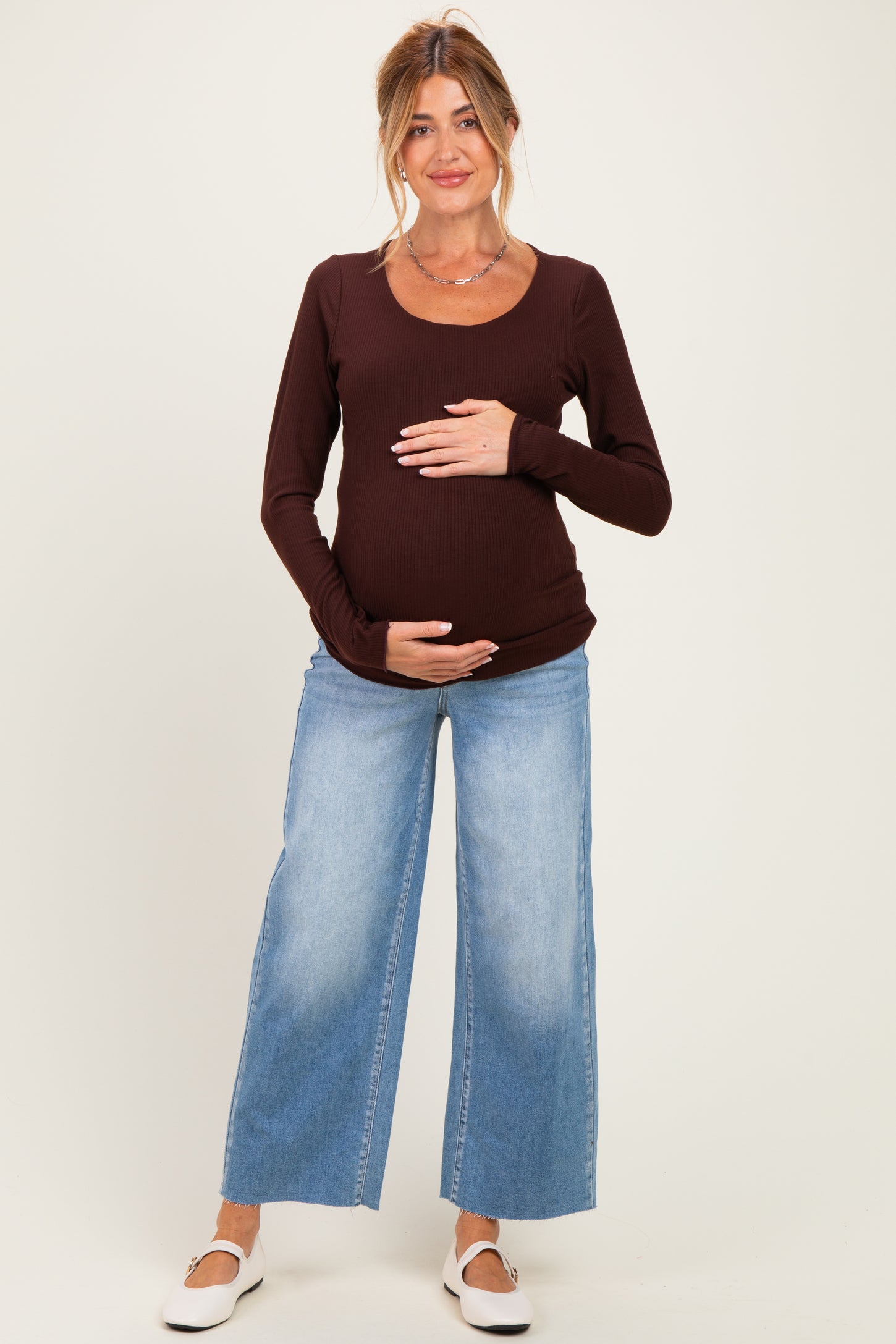 woman wearing a brown long sleeve maternity top and light blue wide leg jeans, holding her baby bump