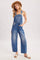 woman wearing a light wash denim overall with wide legs and a hat, standing with hands in pockets