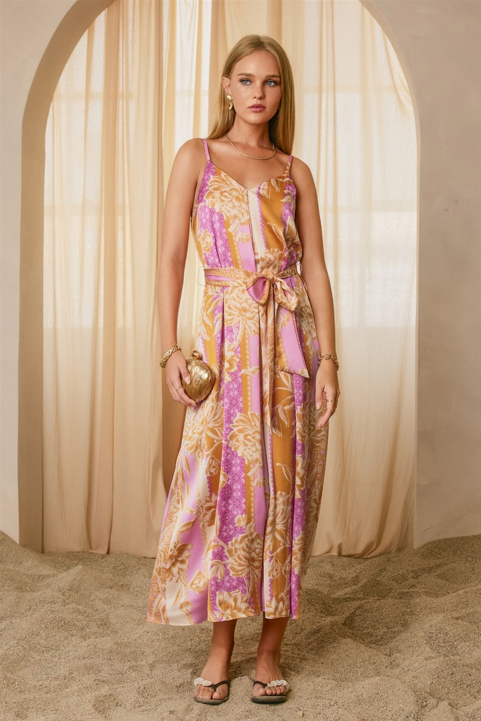 woman wearing a colorful floral maxi dress with a tie waist and spaghetti straps, holding a round clutch bag