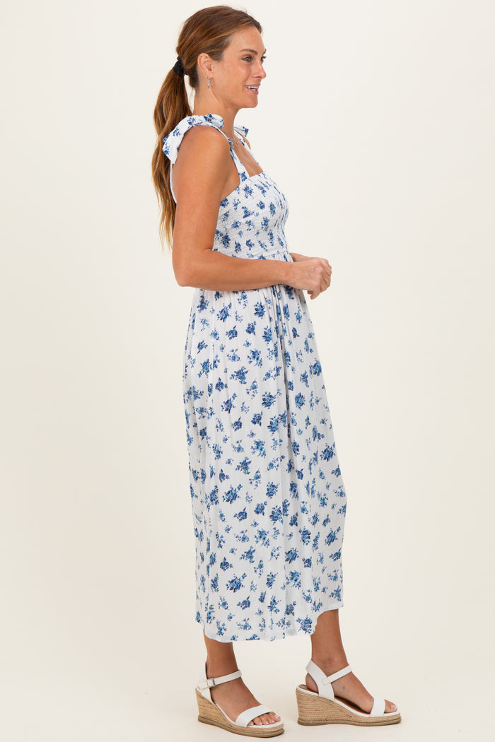woman wearing a white floral midi dress with blue patterns and tied shoulder straps, standing sideways