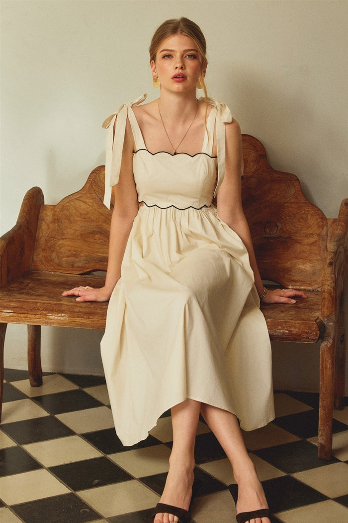 woman wearing a cream midi dress with tie straps and black trim, sitting on a wooden bench