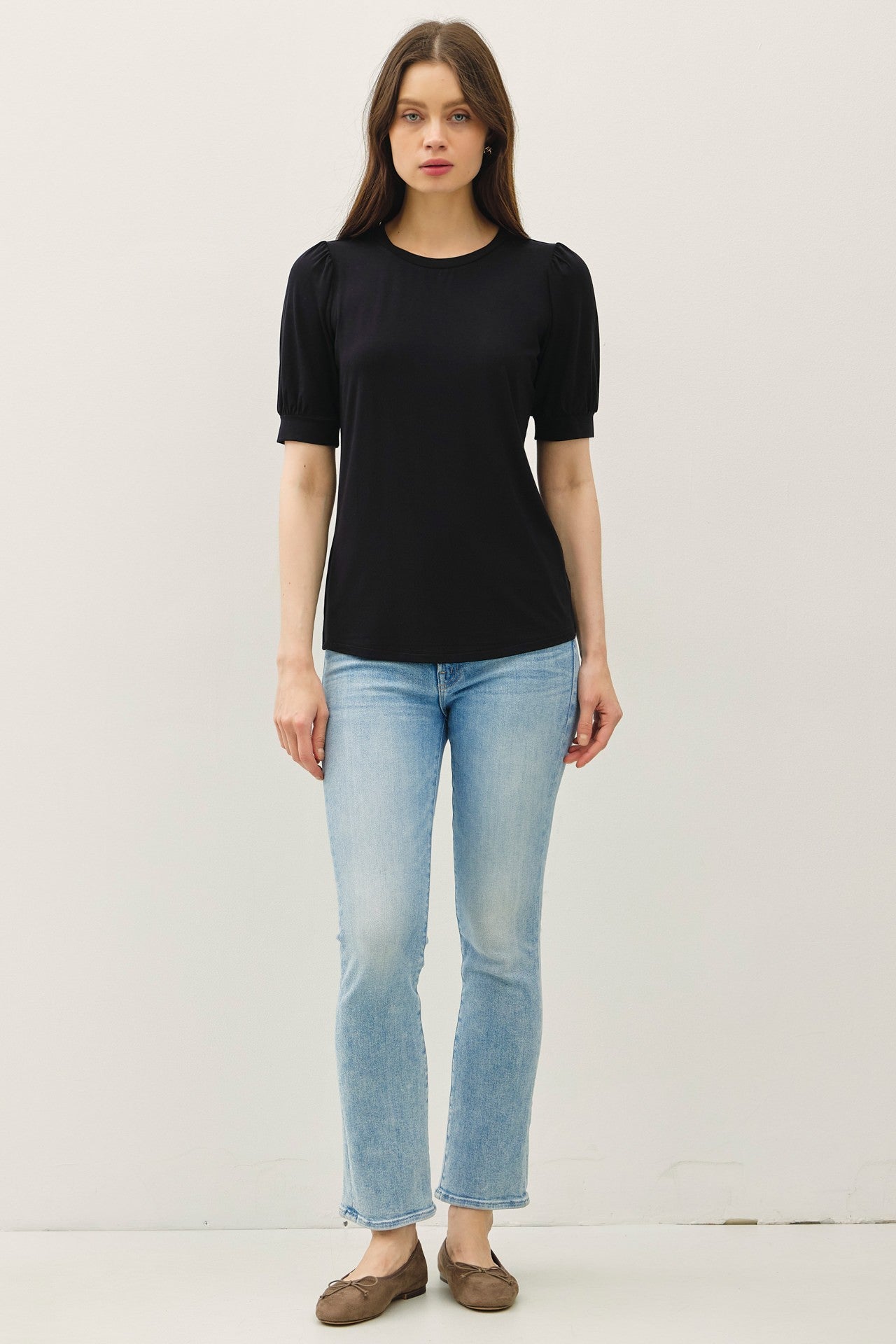woman wearing a black short sleeve top with puff sleeves and light blue flared jeans, standing in a neutral setting