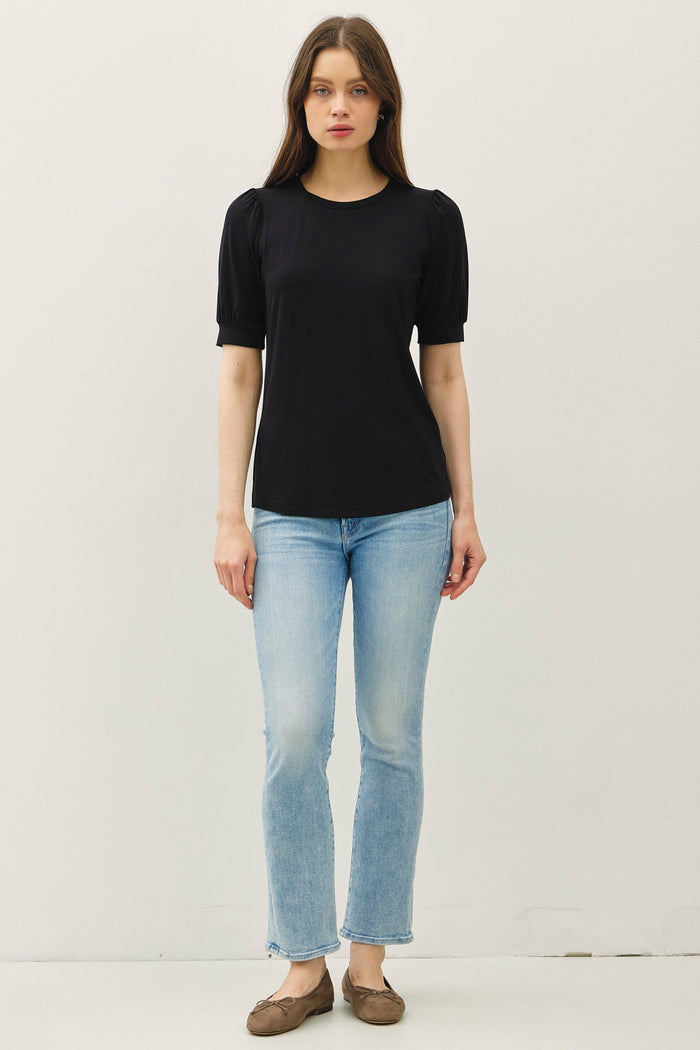 woman wearing a black short sleeve top with puff sleeves and light blue flared jeans, standing in a neutral setting