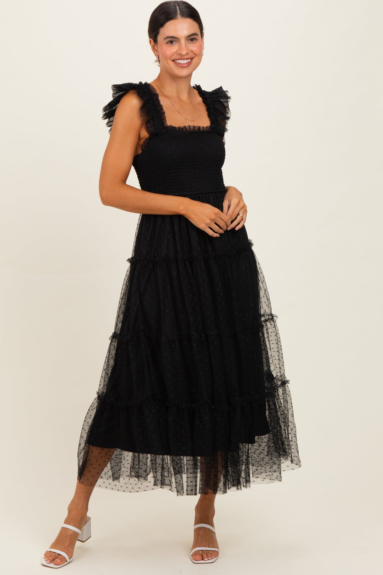 woman wearing a black tiered maxi dress with ruffled straps and sheer overlay, accessorized with white strappy heels