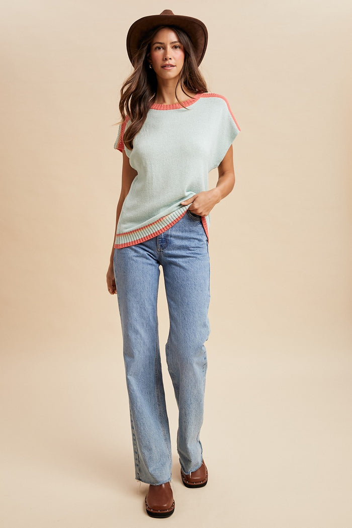 woman wearing a light green short sleeve sweater with coral trim and blue flared jeans, styled with a brown hat