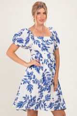 woman wearing a white short sleeve dress with blue leaf print and a square neckline, posing with one hand on her hip