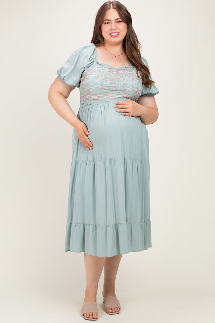 woman wearing a light green maternity plus dress with puff sleeves and embroidered bodice, holding her baby bump