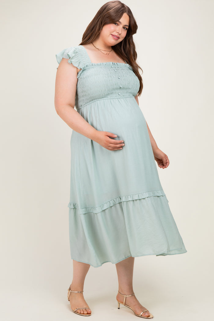 woman wearing a light green maternity plus dress with a smocked bodice and ruffled sleeves, holding her baby bump