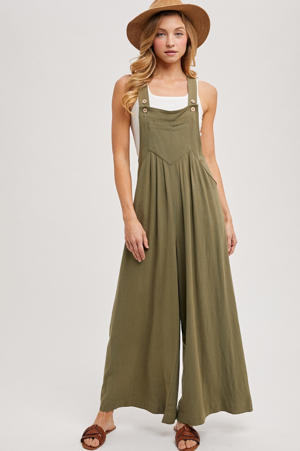 woman wearing an olive green sleeveless jumpsuit with wide legs and buttons, paired with a white tank top and a hat