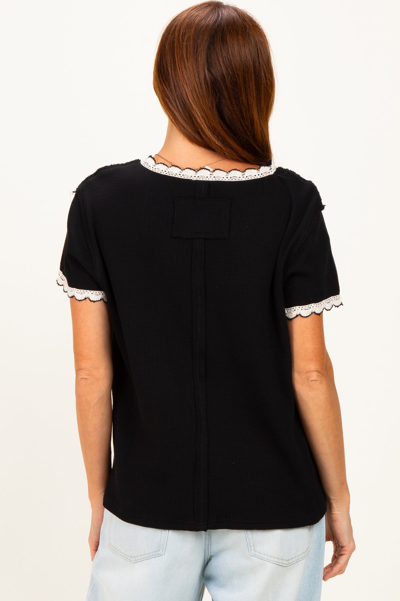 woman wearing a black short sleeve top with scalloped white trim and a relaxed fit, viewed from the back
