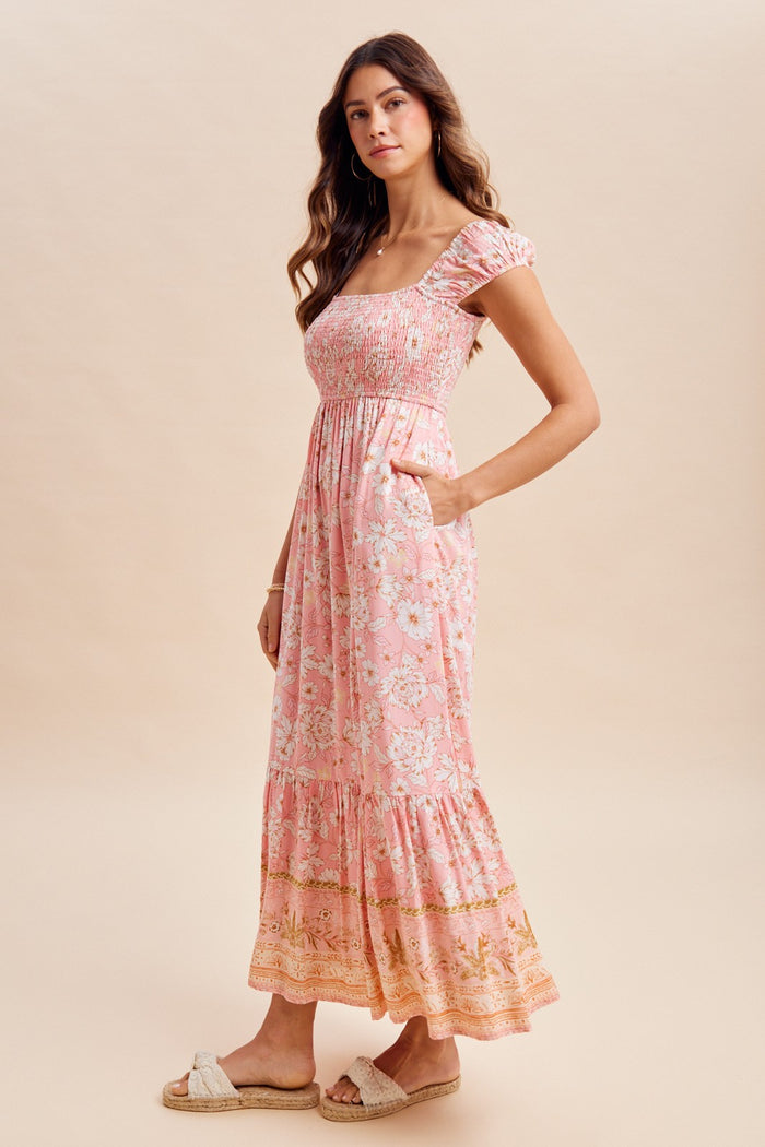 woman wearing a pink floral maxi dress with a square neckline and puff sleeves, featuring a tiered hem and pockets