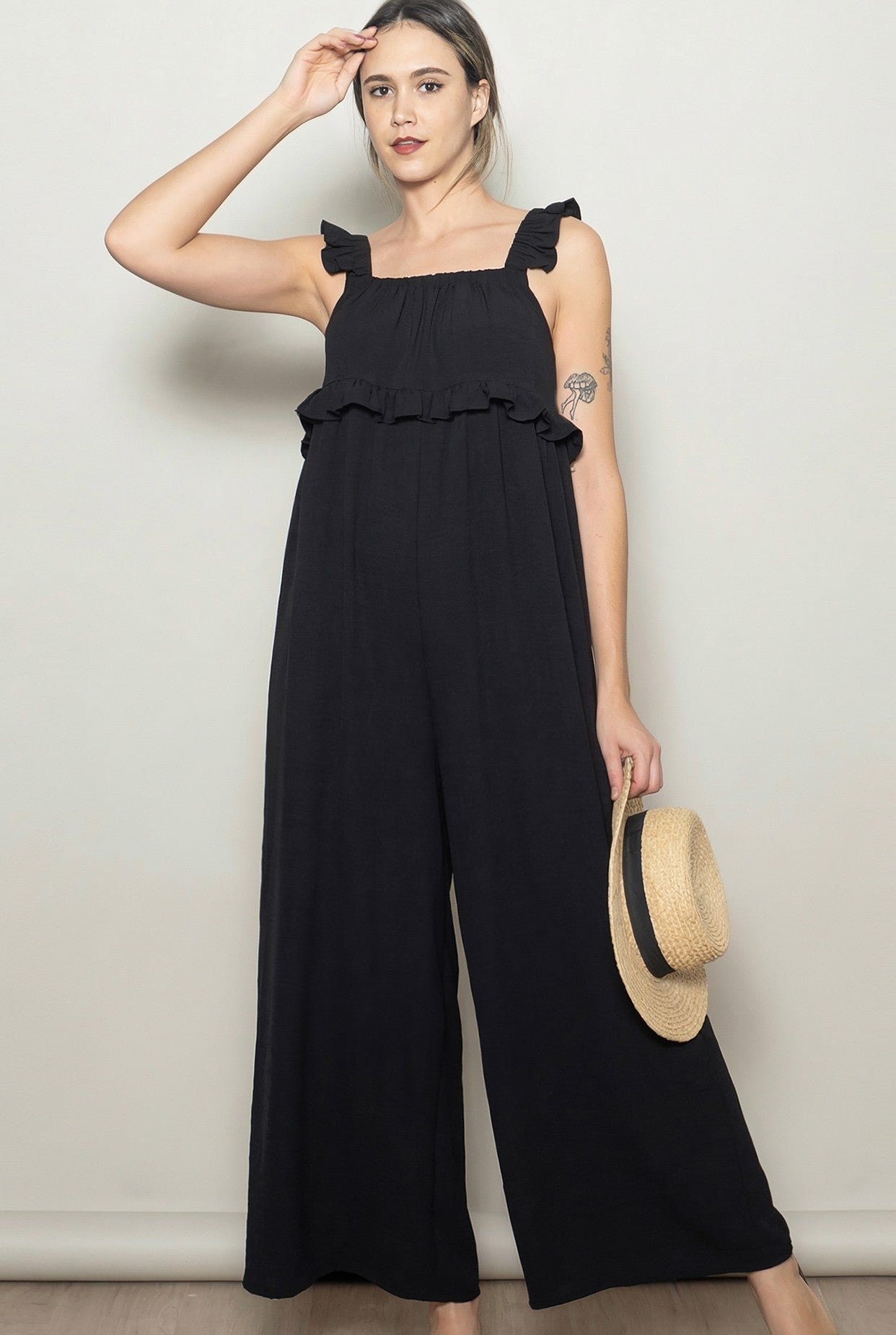 woman wearing a black ruffled strap jumpsuit and holding a straw hat