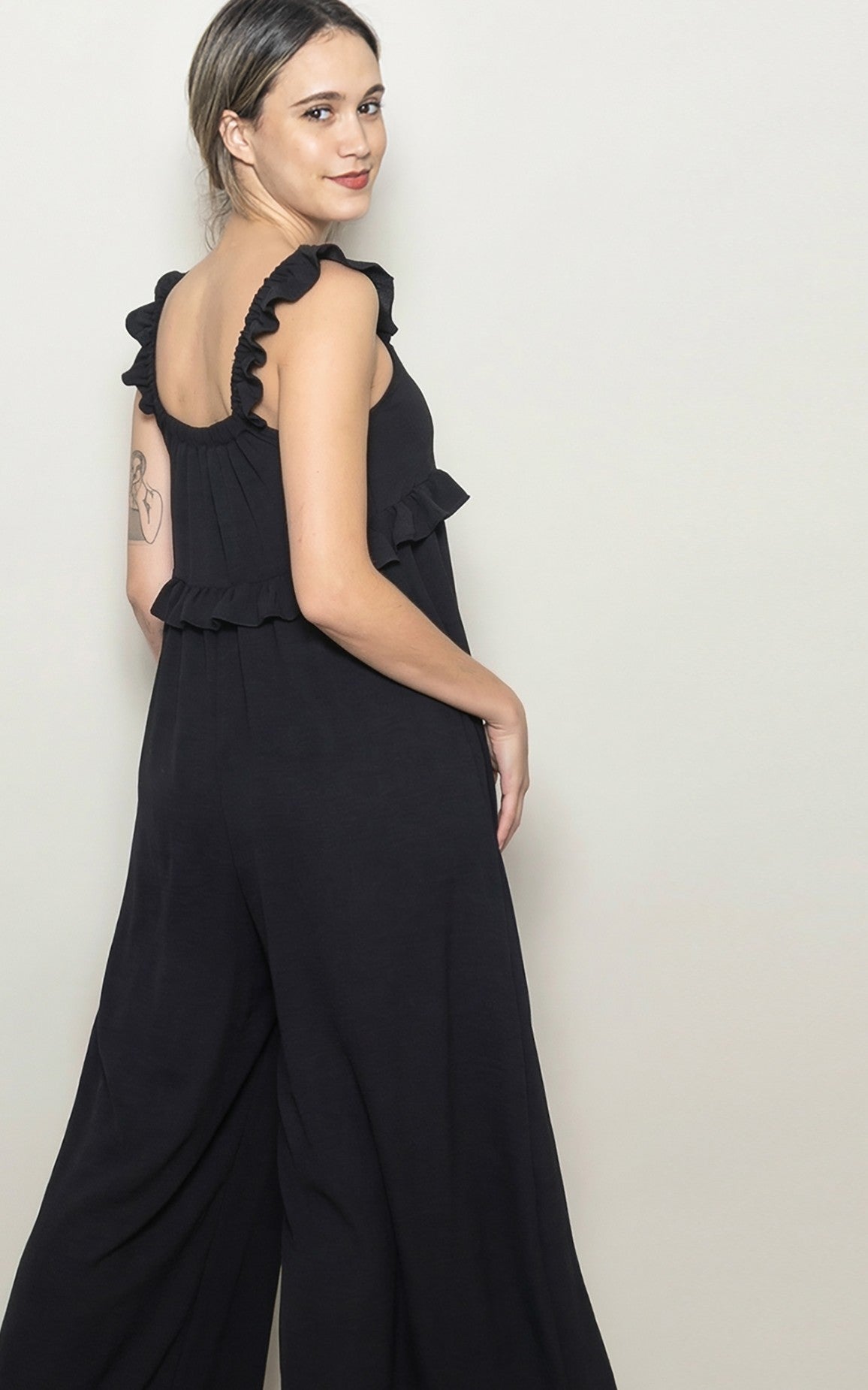 woman wearing a black jumpsuit with ruffled straps and a flared leg, posed sideways against a neutral background
