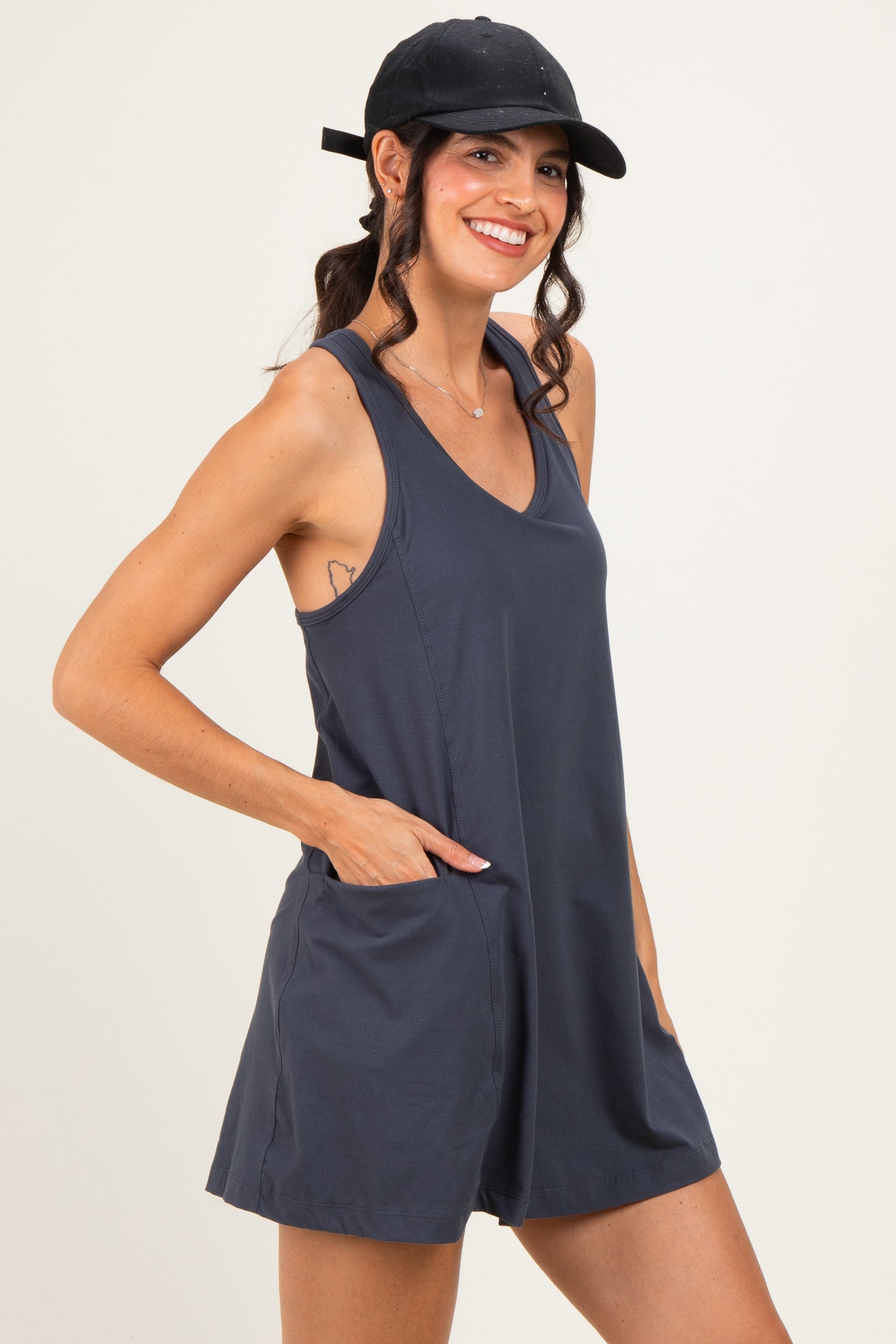 woman wearing a dark gray sleeveless romper with pockets, smiling and wearing a black cap
