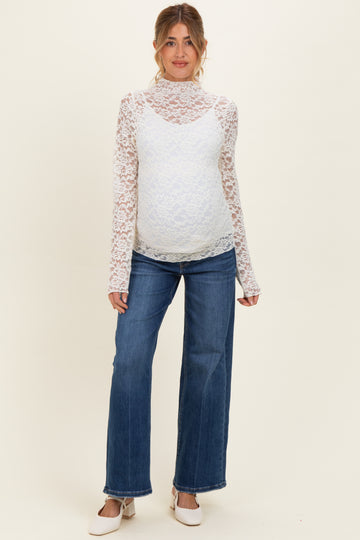 woman wearing a white lace maternity top with long sleeves and blue wide-leg maternity jeans, showcasing her baby bump
