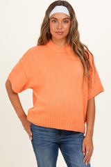 woman wearing an orange oversized maternity sweater with short sleeves and a ribbed hemline, styled with blue jeans