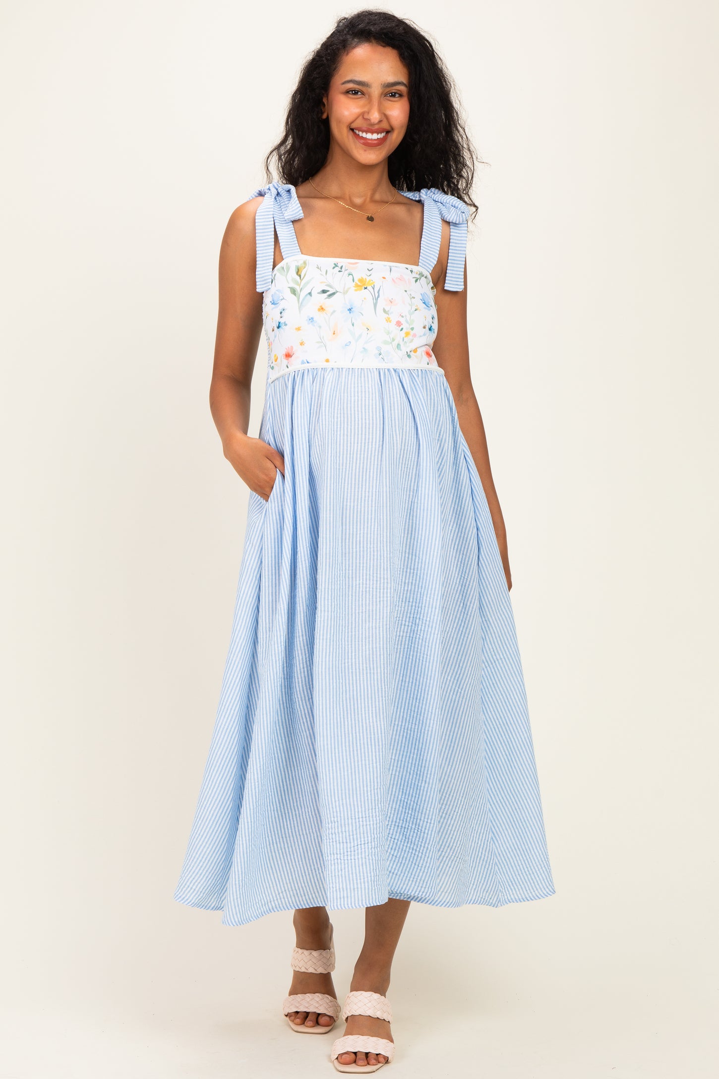 woman wearing a light blue striped maternity dress with floral embroidery and tie straps, posing with hands in pockets