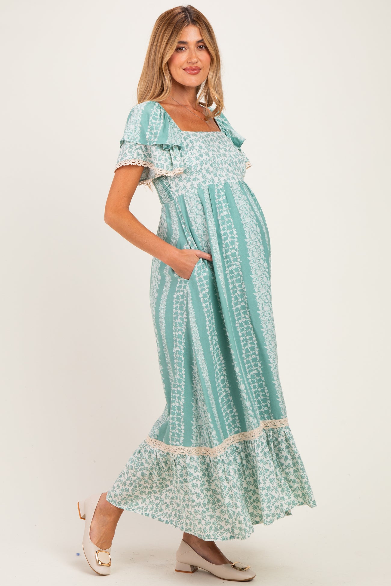 woman wearing a light teal floral print maternity dress with ruffled sleeves and pockets, showcasing her baby bump