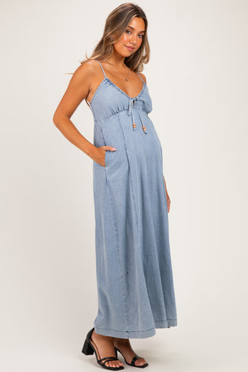 woman wearing a light blue denim maternity maxi dress with adjustable straps and pockets, showcasing her baby bump