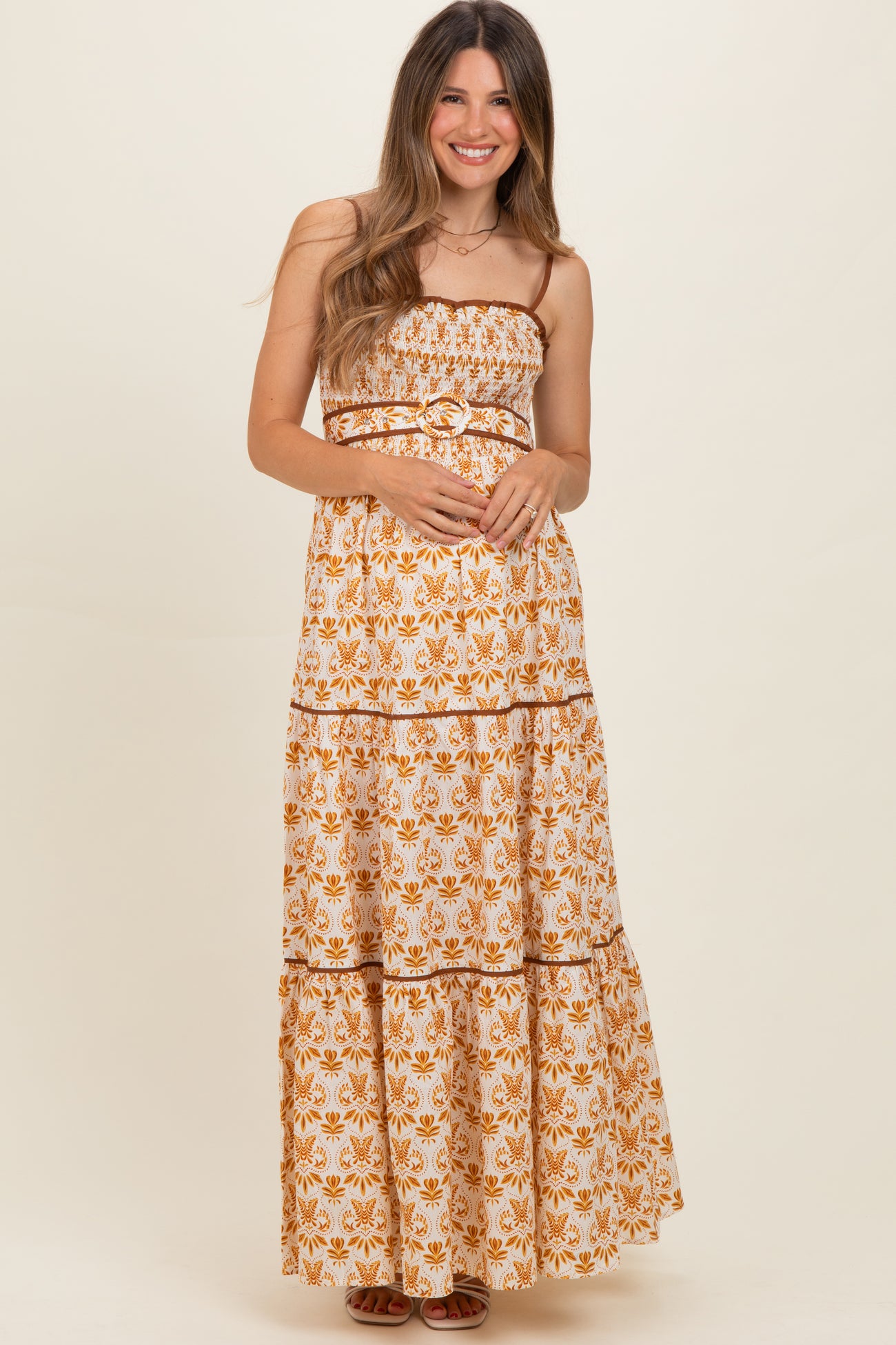 woman wearing a maternity maxi dress with a white and orange floral pattern and a belted waist, smiling and holding her baby bump