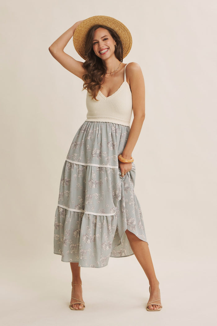 woman wearing a cream knitted tank top and floral tiered midi skirt, smiling and holding a straw hat