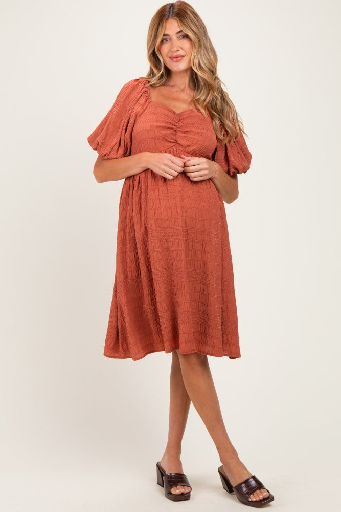 woman wearing a rust-colored maternity dress with puff sleeves and a gathered bust, standing in a relaxed pose