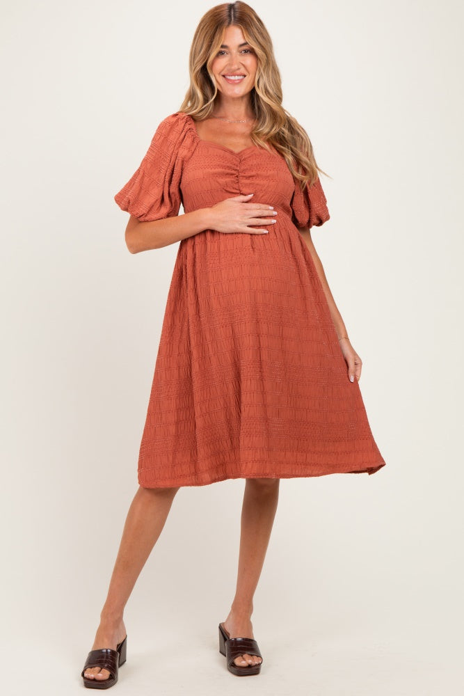 woman wearing a rust-colored maternity dress with puff sleeves and a textured fabric, holding her baby bump
