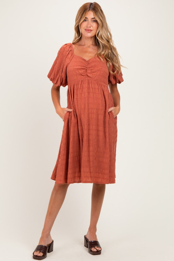 woman wearing a rust colored maternity dress with puff sleeves and pockets, featuring a gathered bust and relaxed fit