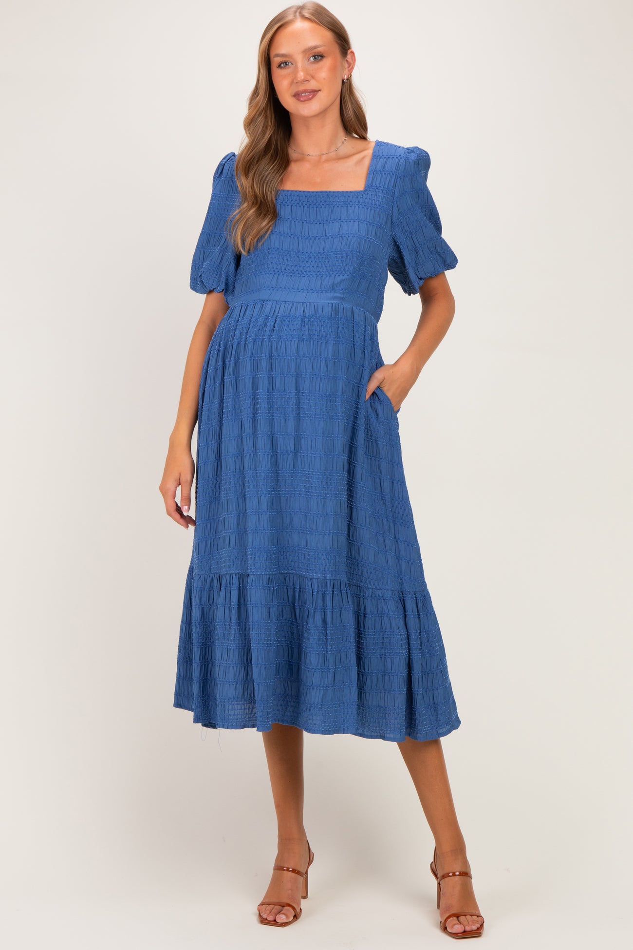 woman wearing a blue maternity dress with puff sleeves and tiered skirt, standing with hands in pockets and showing baby bump