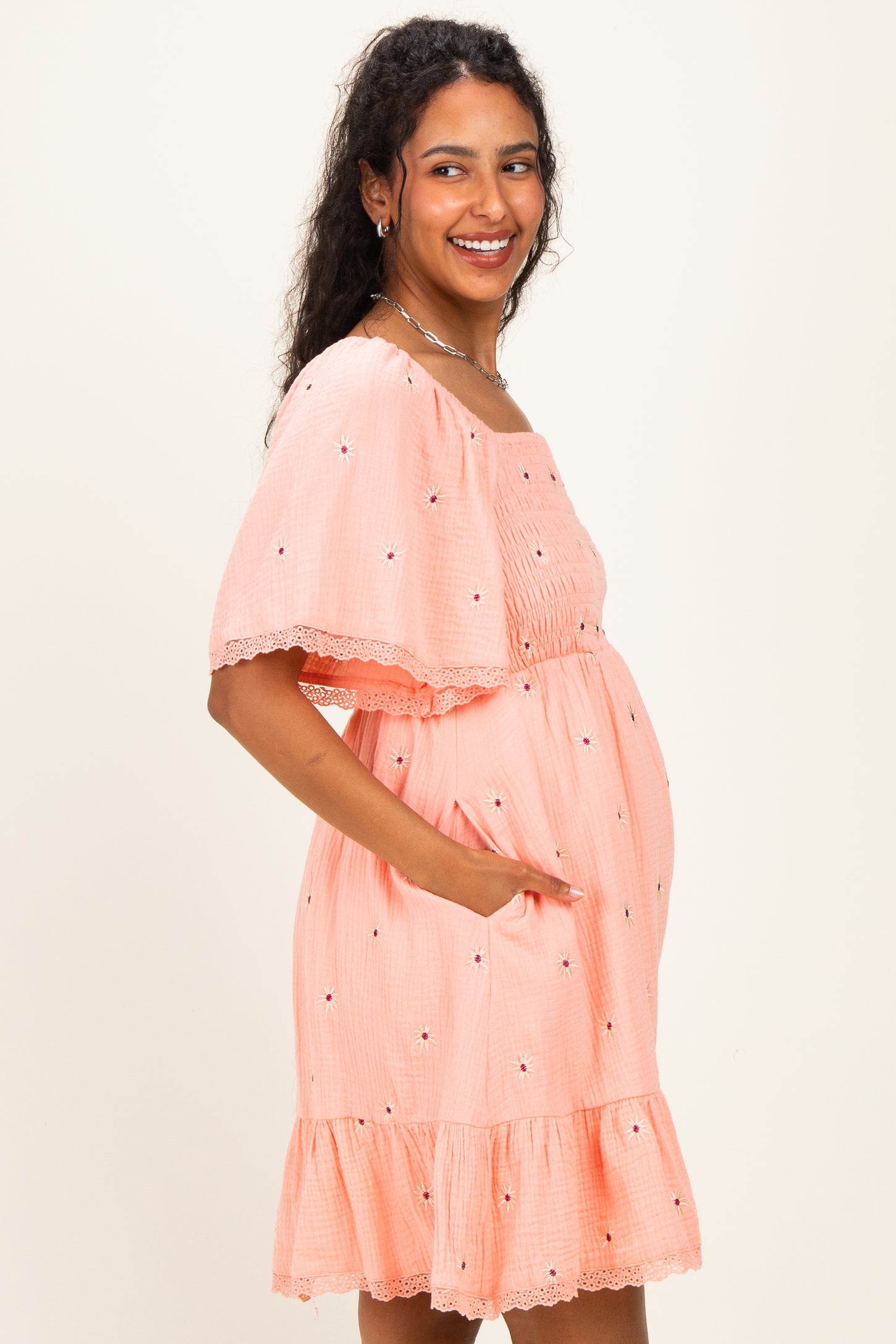 woman wearing a coral maternity dress with a ruffled hem, short sleeves, and floral embroidery, showcasing her baby bump