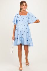 woman wearing a light blue maternity dress with floral embroidery and short puff sleeves, holding a white handbag