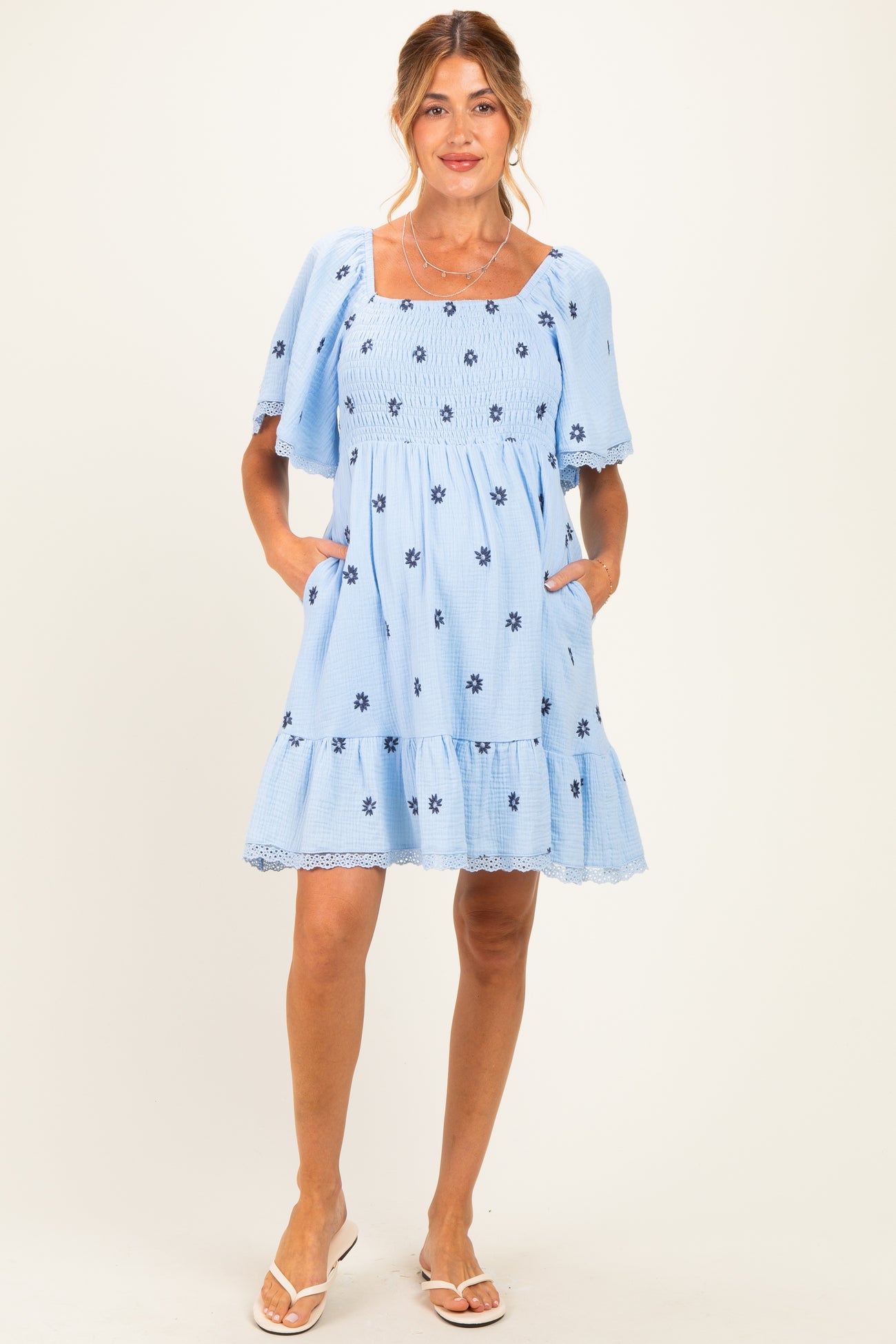 woman wearing a light blue maternity dress with floral embroidery and short flutter sleeves, featuring pockets