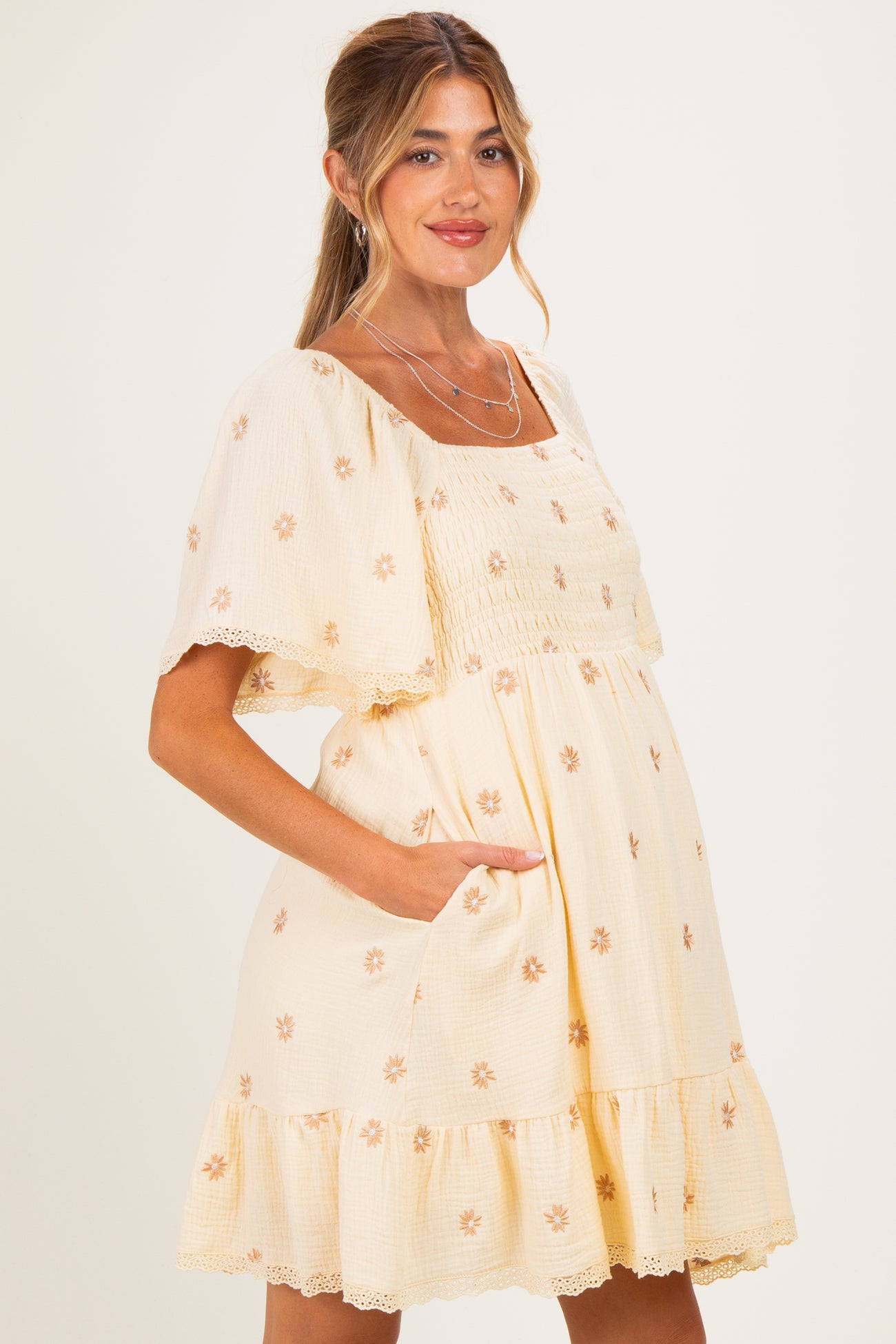 woman wearing a cream maternity dress with floral embroidery and ruffled hem, showing her baby bump and smiling