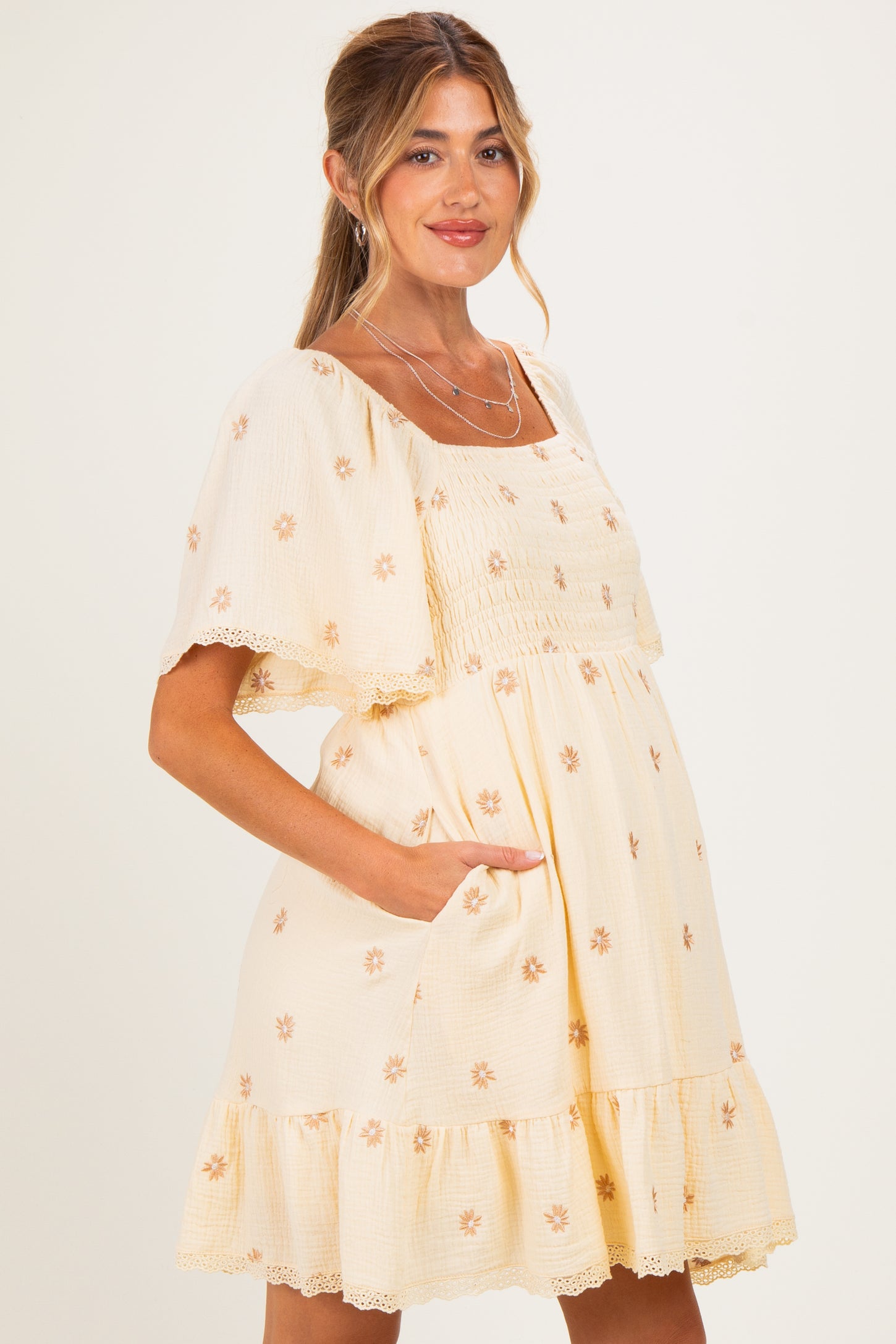 woman wearing a cream maternity dress with floral embroidery and ruffled hem, showing her baby bump and smiling