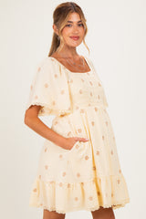 woman wearing a cream maternity dress with floral embroidery and ruffled hem, showing her baby bump and smiling
