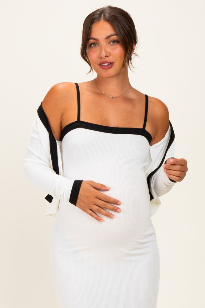 woman wearing a white ribbed maternity dress with black trim and a matching cardigan, cradling her baby bump