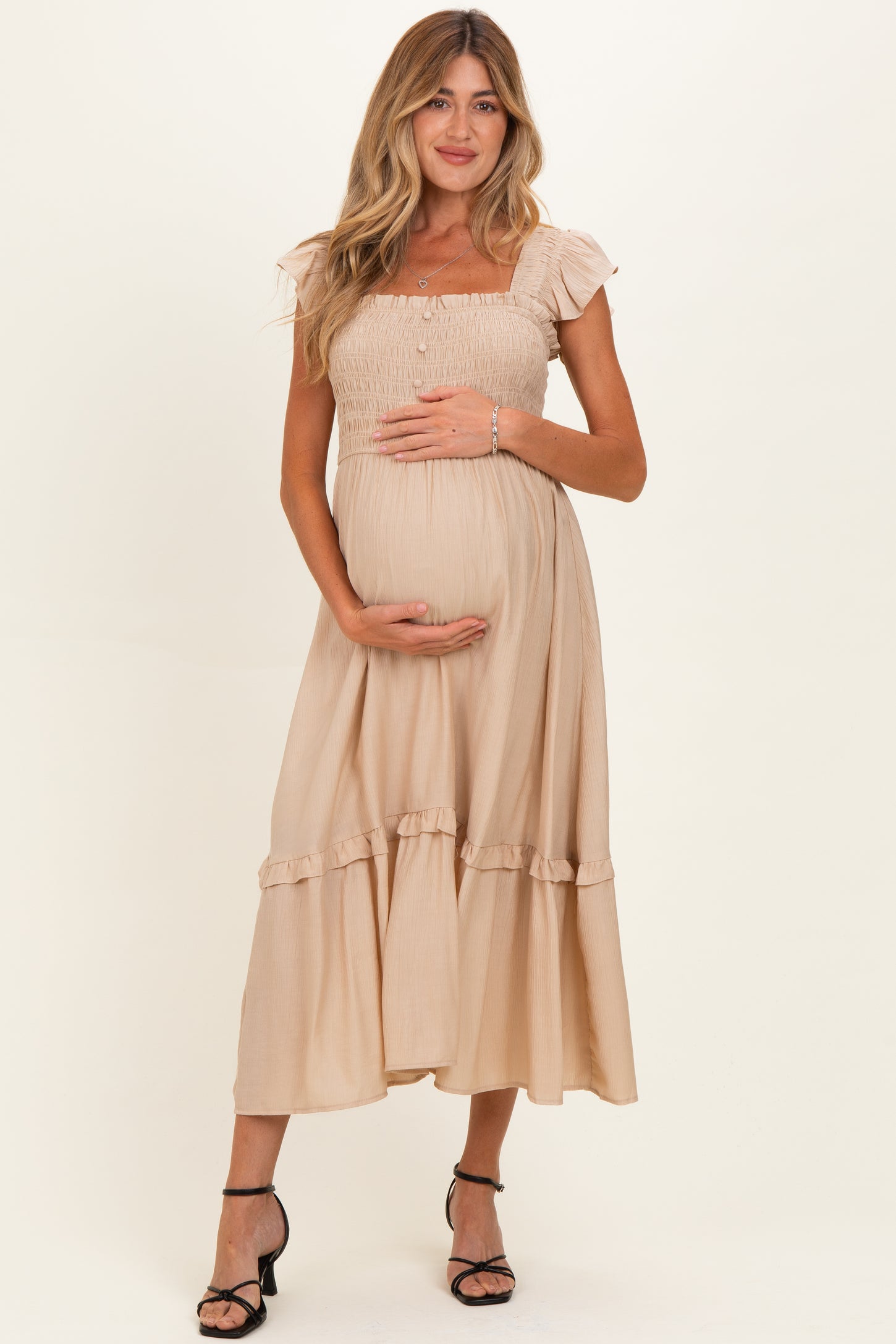 woman wearing a beige maternity dress with flutter sleeves and ruffle hem, holding her baby bump