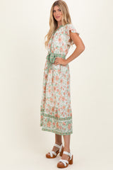 woman wearing a floral maxi dress with a tie waist and green accents, paired with white block heel sandals