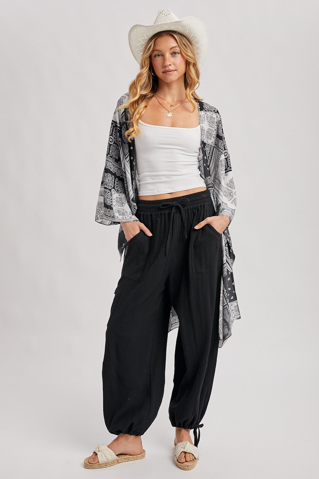woman wearing a white tank top, black patterned kimono, and black loose-fitting pants with pockets, styled with a wide-brimmed hat