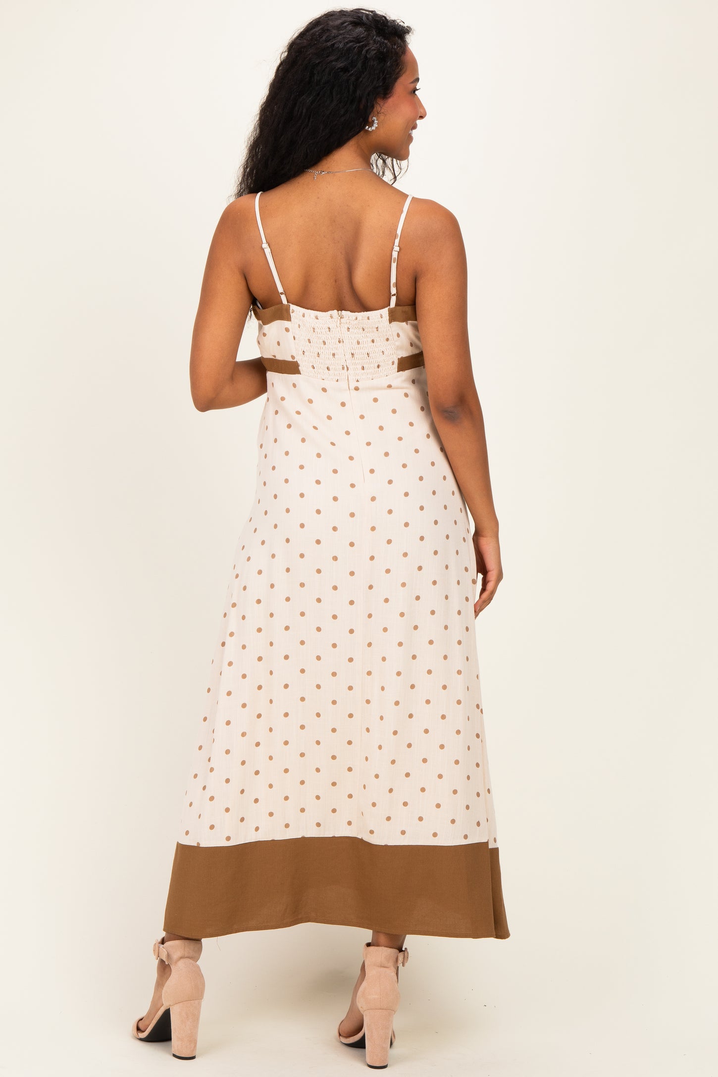woman wearing a cream maternity maxi dress with polka dots and a brown hem, showing her back and smocked bodice