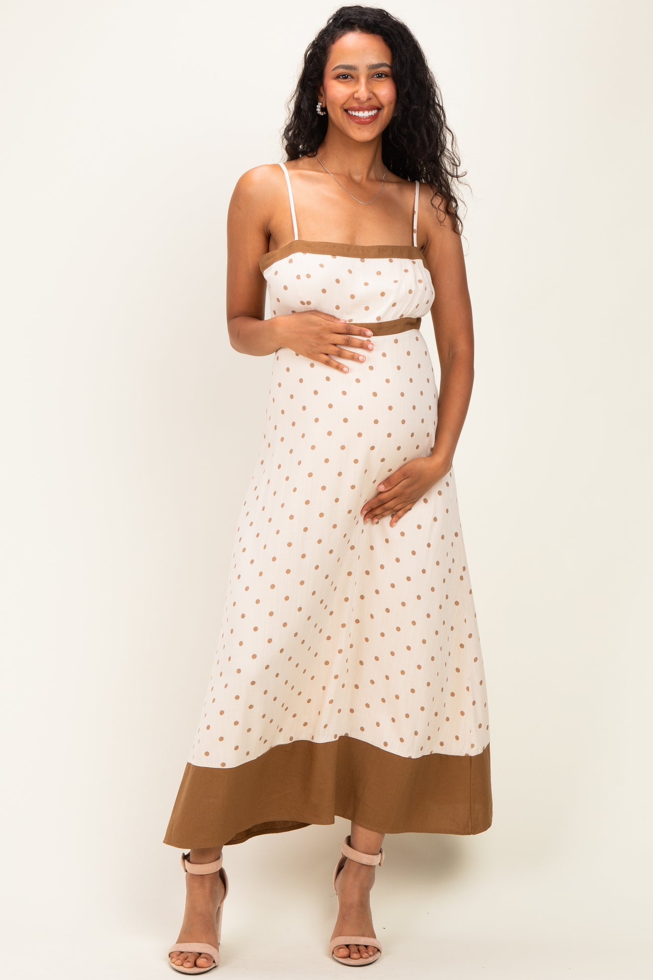 woman wearing a cream and brown polka dot maternity maxi dress with a baby bump and spaghetti straps