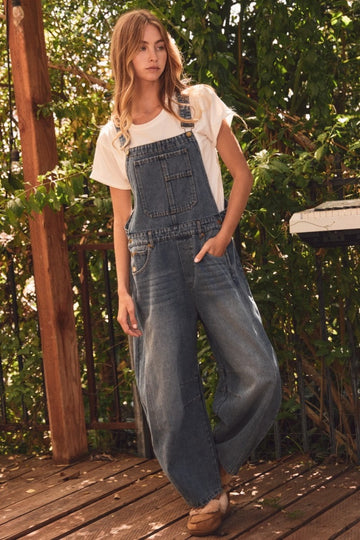 woman wearing a light blue denim overall with a white t-shirt and standing in a natural outdoor setting