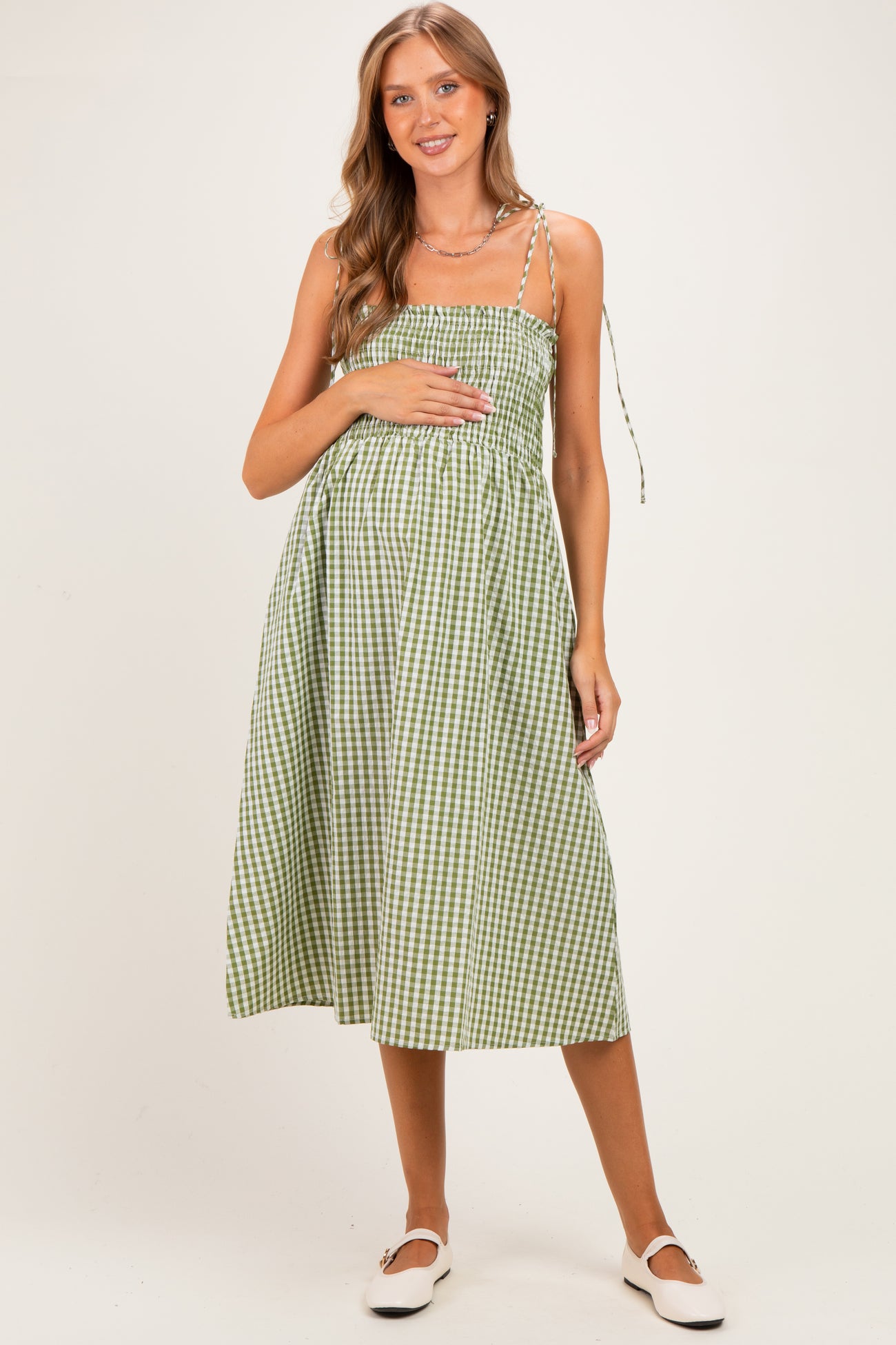 woman wearing a green and white gingham maternity dress with adjustable straps and holding her baby bump