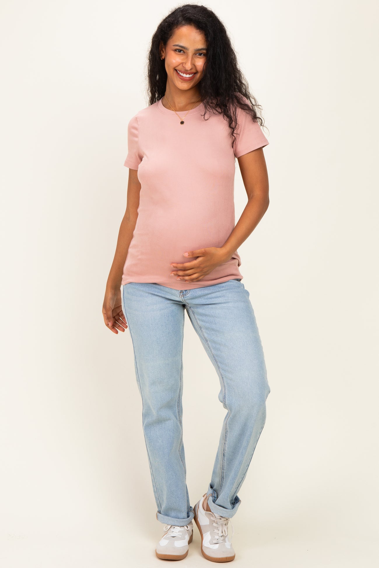 woman wearing a light pink maternity top and blue jeans, smiling while holding her baby bump