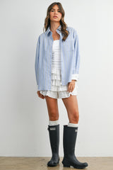 woman wearing a light blue striped oversized button-up shirt, white tank top, and ruffled shorts with black rain boots
